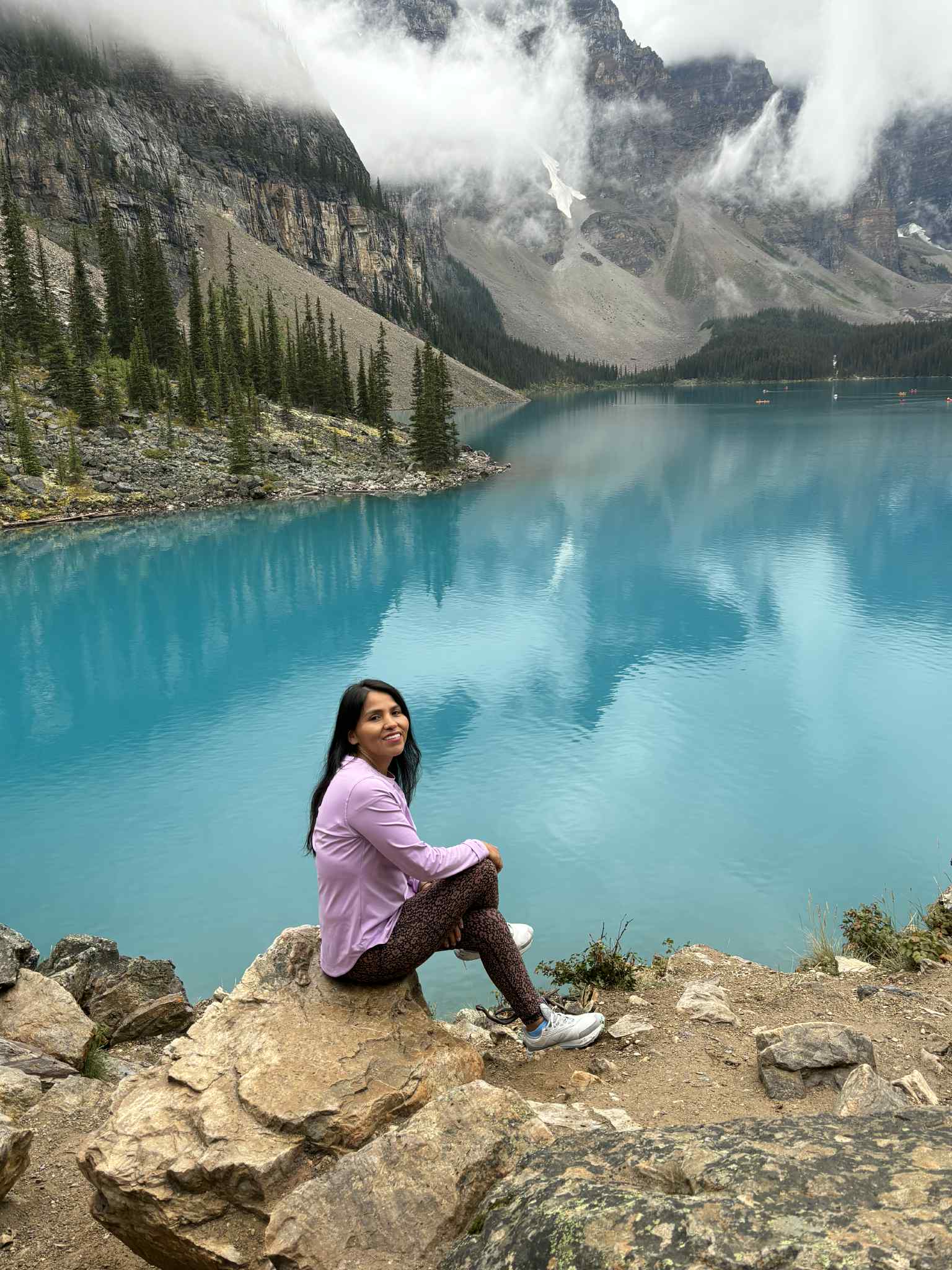 Femme en mauve assise sur un rocher devant un lac turquoise et des montagnes brumeuses.