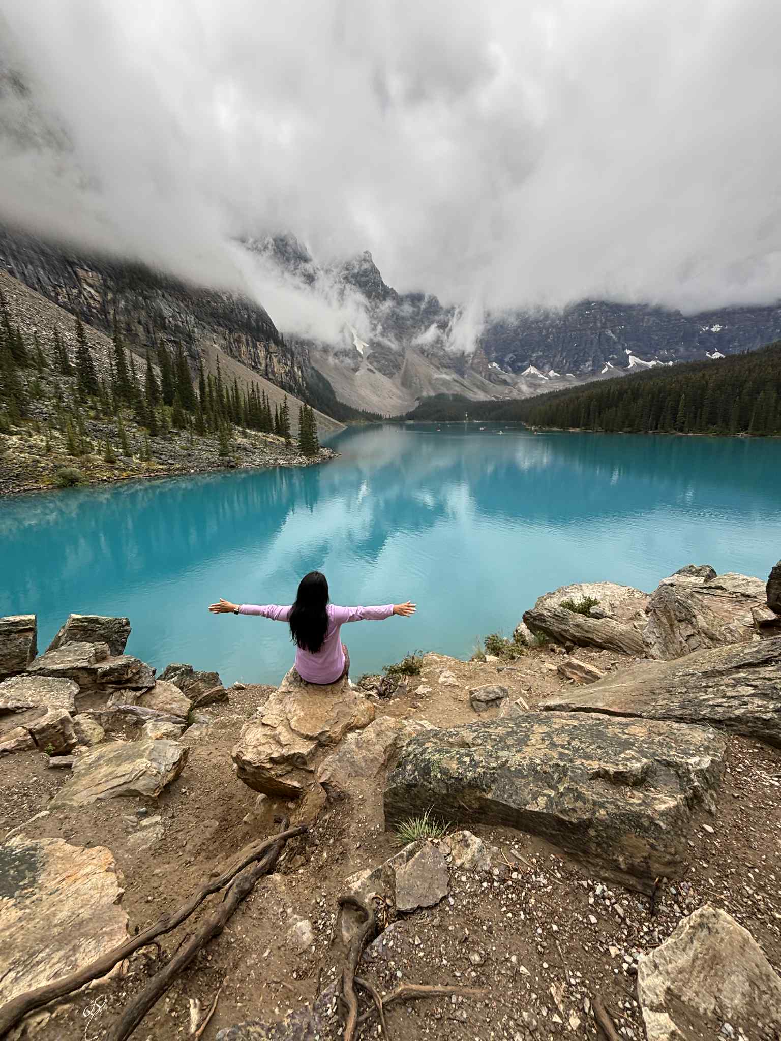 Femme assise sur un rocher, bras ouverts devant le lac Moraine turquoise et montagnes embrumées.