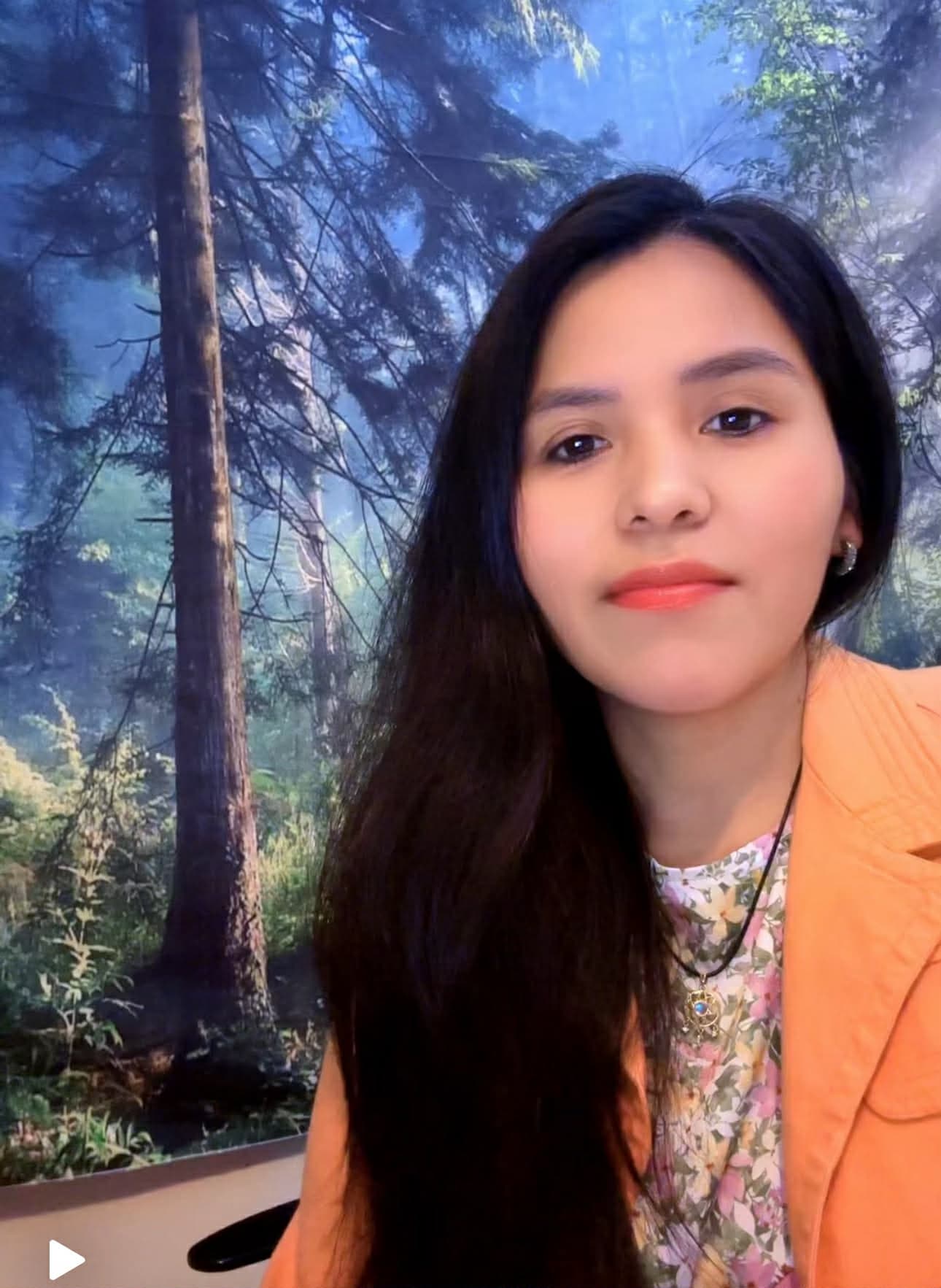 Femme aux longs cheveux noirs portant un veston orange devant un décor de forêt boréale.